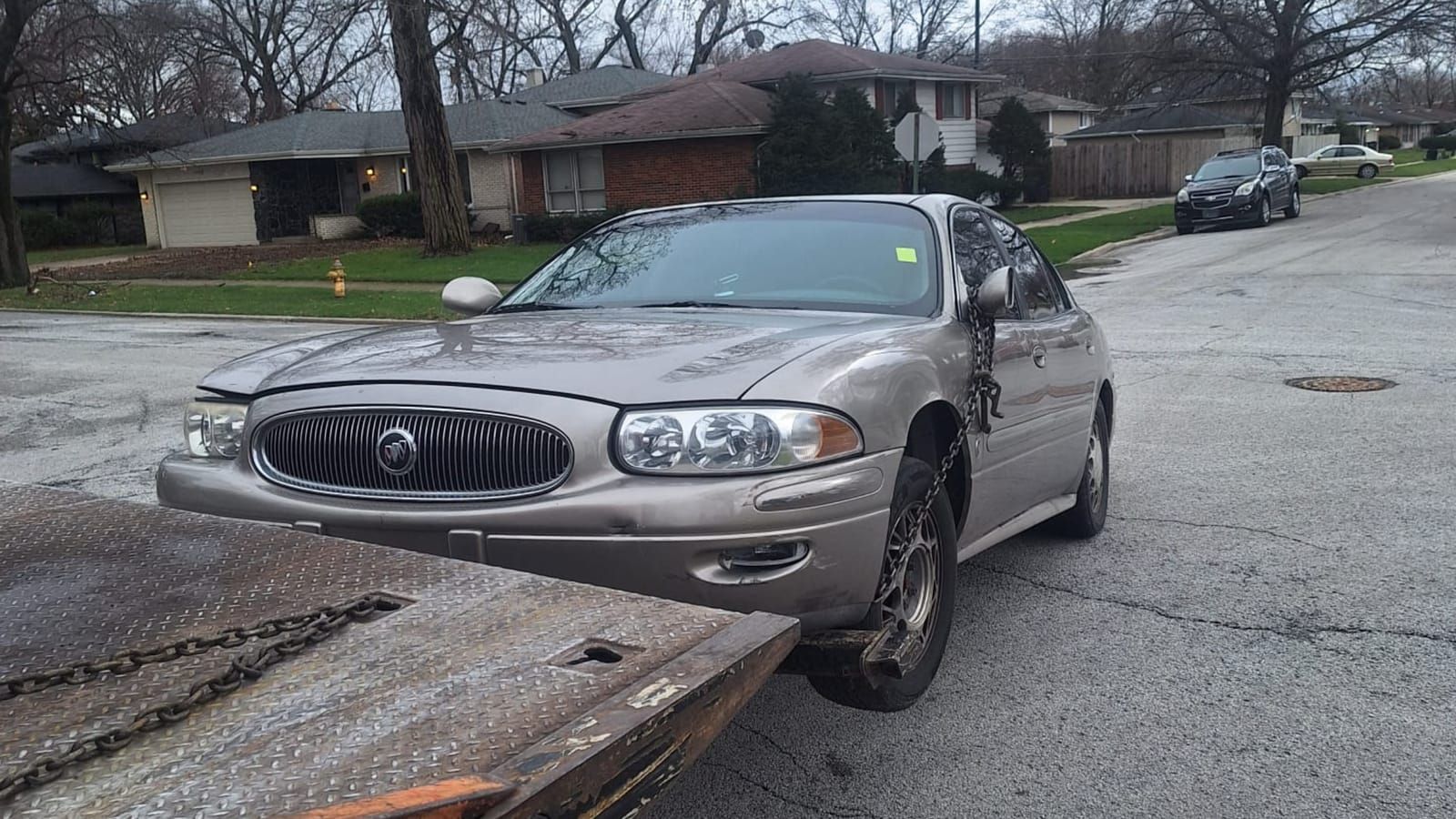 Champagne Buick LeSabre being chained to the tilted deck of a flatbed on a residential street