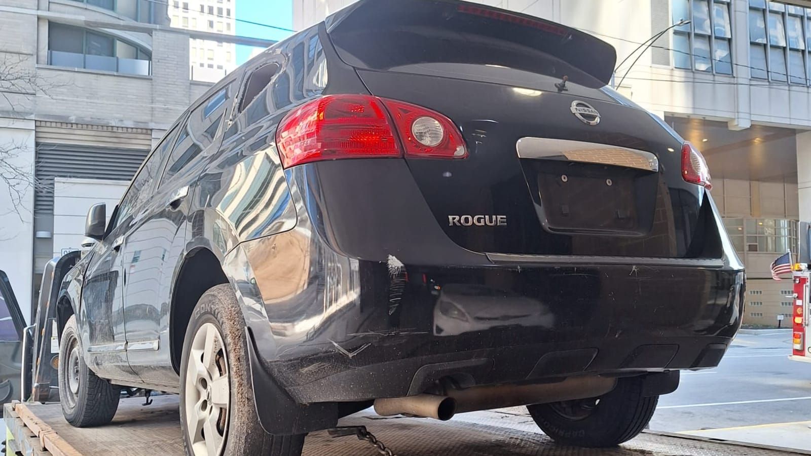 Dark blue Nissan Rogue with its rear end pushed in, strapped to a flatbed outside a brick apartment building