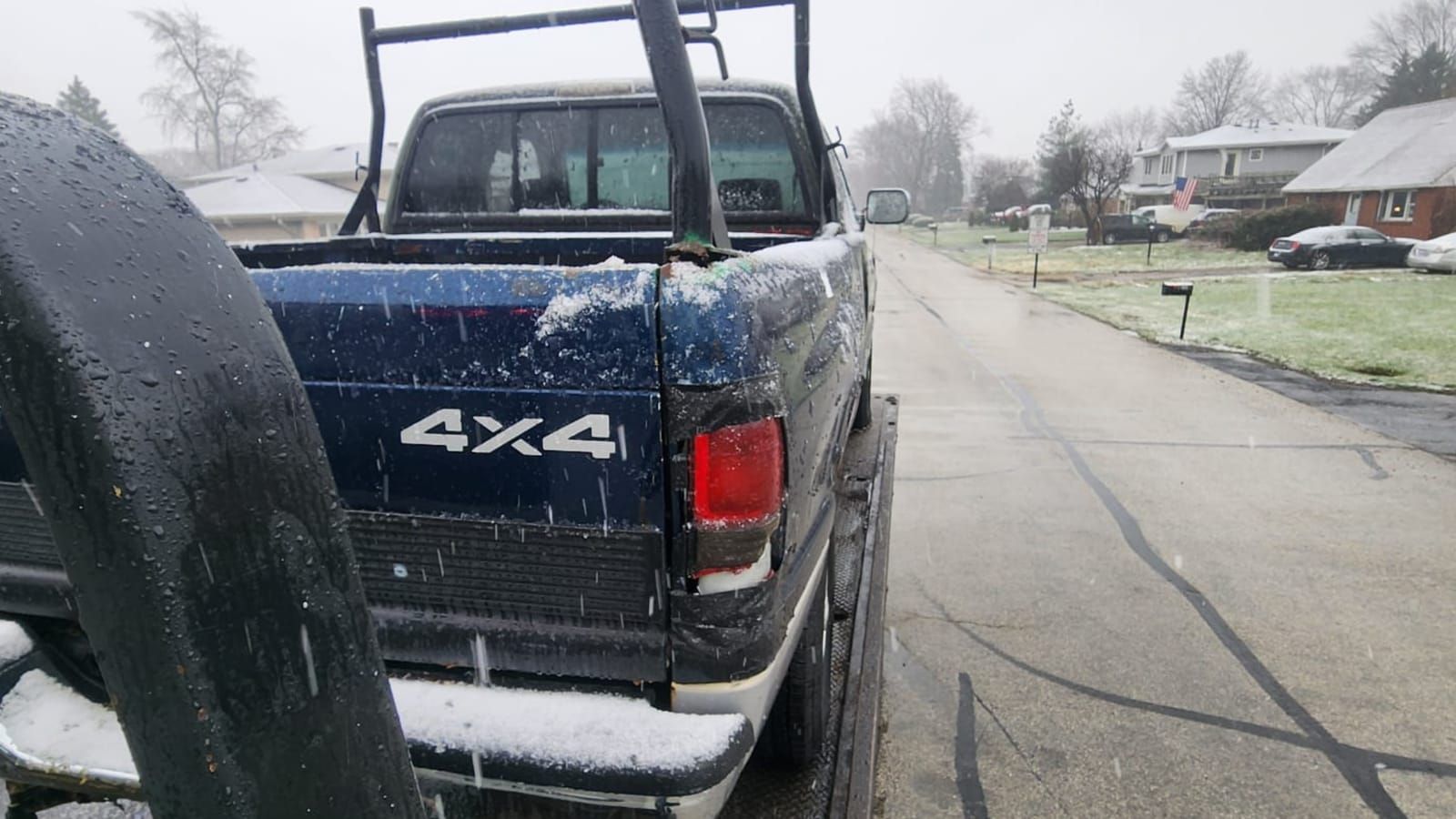White work pickup with a ladder rack, strapped down on a flatbed as snow falls on a suburban street